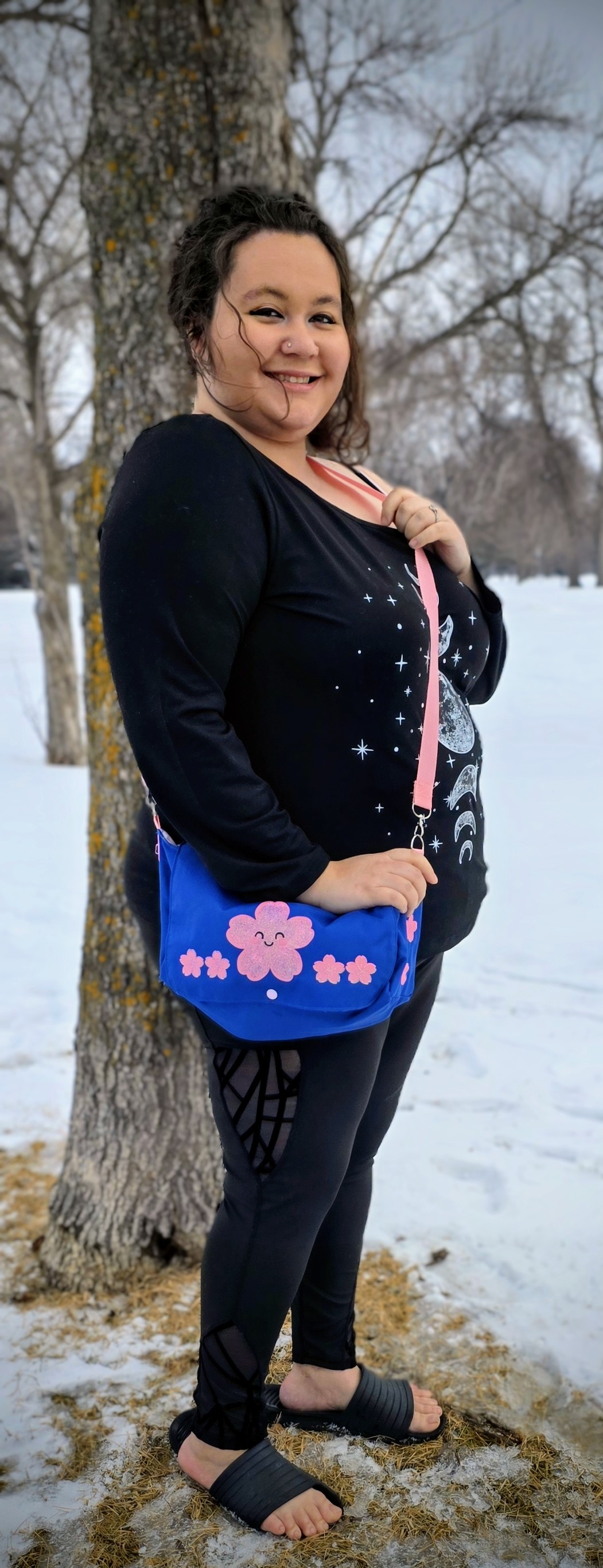 a beautiful woman in black showing off a royal blue diaper purse with a pink strap and pink kawaii cherry blossoms ironed on the front. in the background of the image are snow and trees