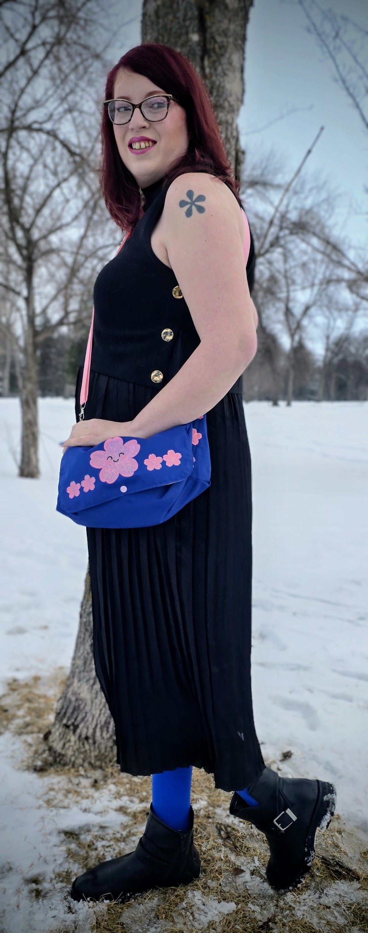 a women in a black dress looking behind her wearing a royal blue diaper purse with a pink strap and pink kawaii cherry blossoms ironed on the front of it. the woman has a black asterisk tattooed on her shoulder and is wearing glasses.