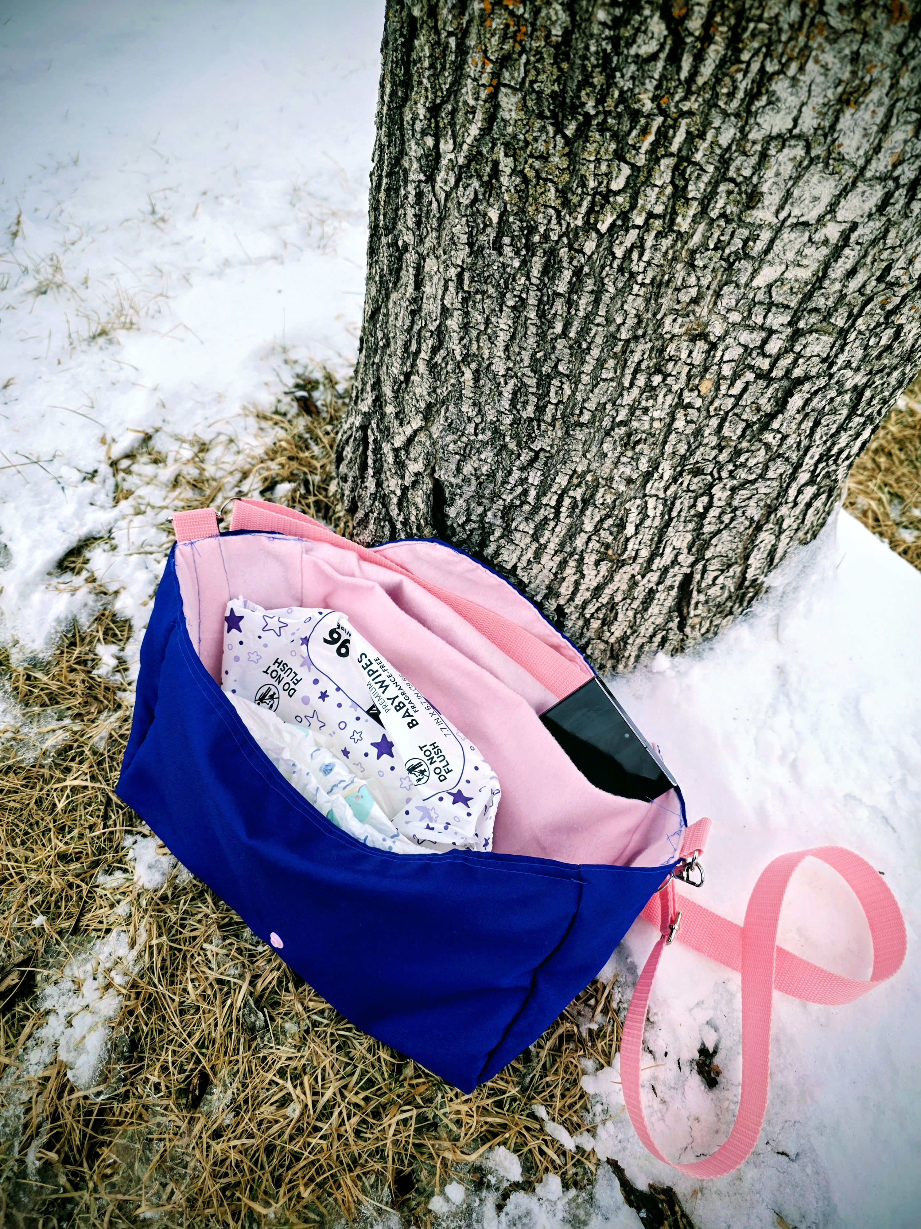 open royal blue purse lined with light pink flannel with a flannel pocket and pink strap sitting up against a tree against the snow. the purse contains a cell phone, a package of wipes, and two diapers