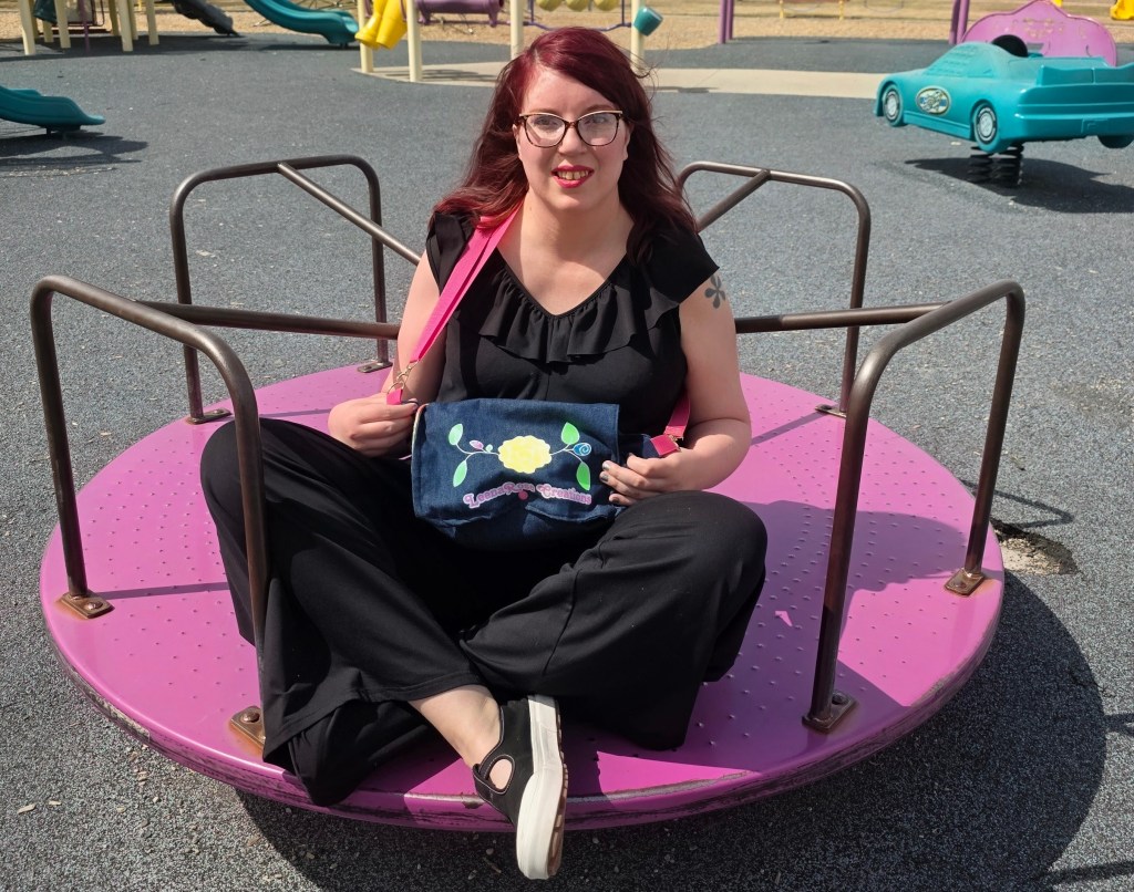 A women on a merry go round in a black jumpsuit and black shoes, wearing a purse with the LeenaRose Creations logo on it. The Purse is denim with a pink strap.