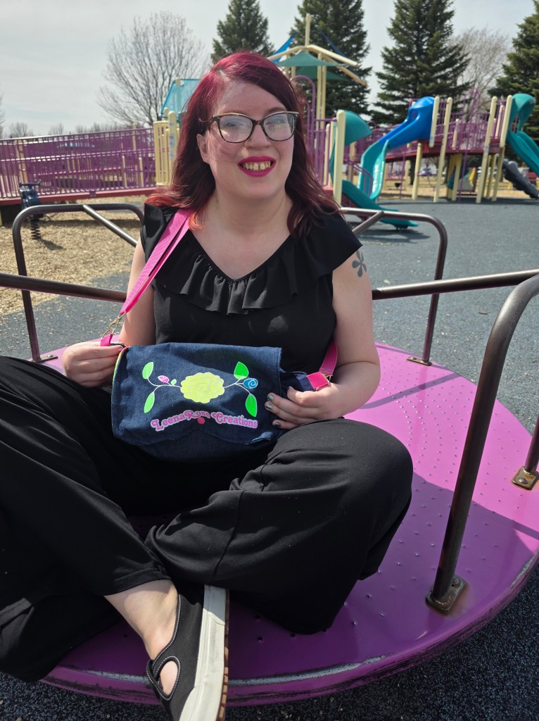 A women on a merry go round in a black jumpsuit and black shoes, wearing a purse with the LeenaRose Creations logo on it. The Purse is denim with a pink strap.