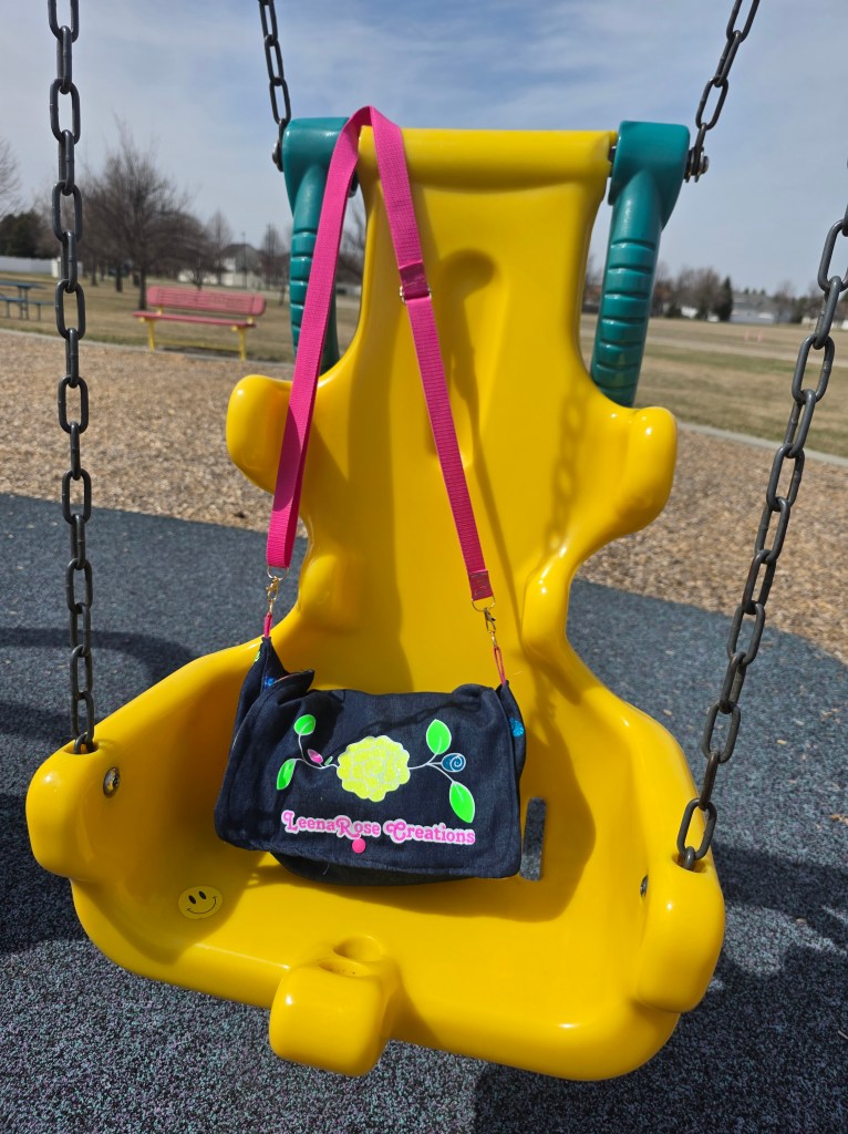 A denim purse with the LeenaRose Creations logo on the front with a pink strap, sitting on an accessible swing that's yellow