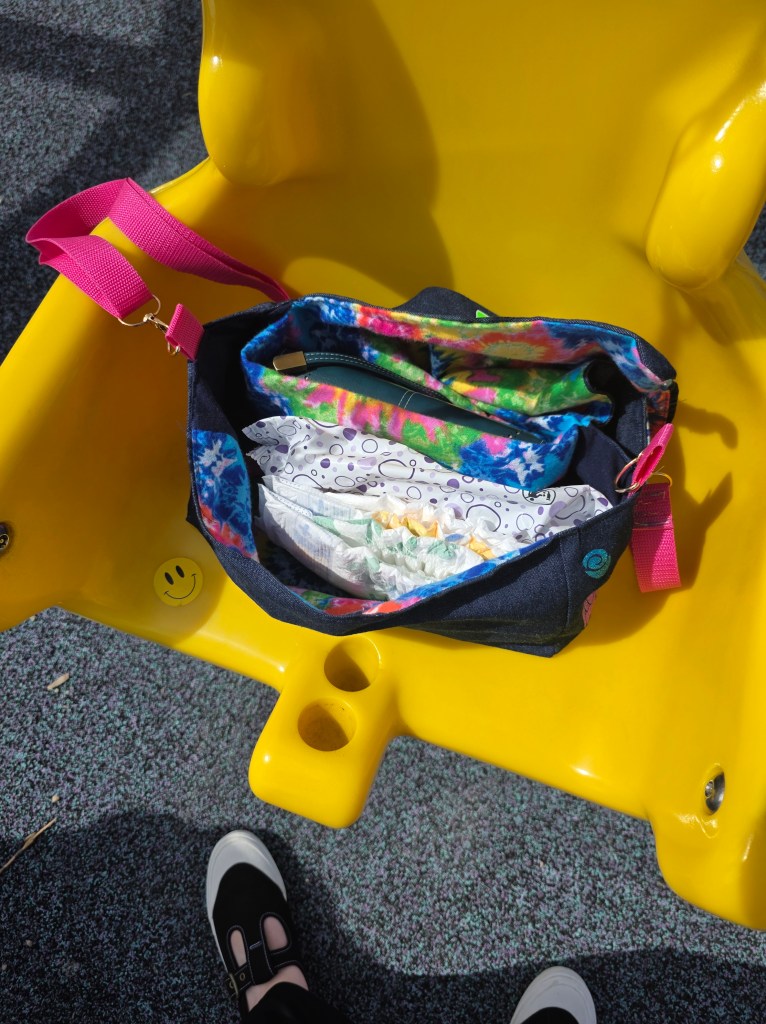 A denim purse sitting on an accessible swing, open to reveal a pocket. The purse contains a wallet, a packet of baby wipes, and two diapers