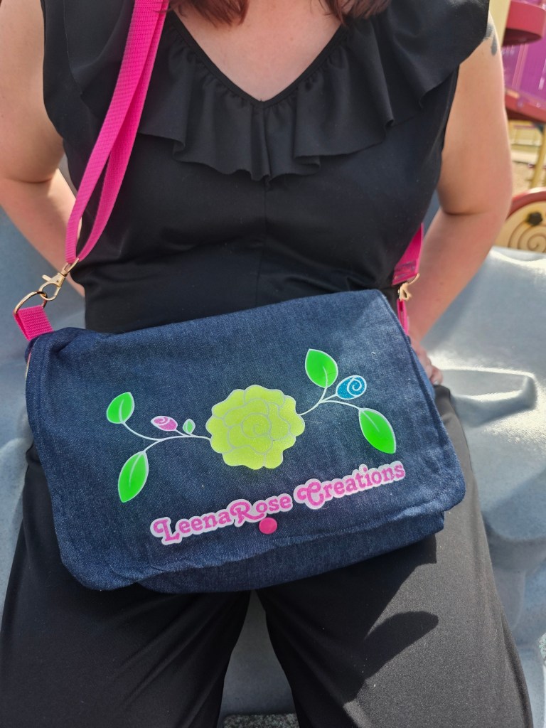 a close up of a torso of a women in a black jumpsuit with a ruffled collar, holding a denim purse with the LeenaRose Creations logo with a pink strap barely visible