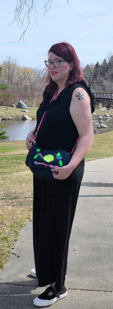 A women in a black jumpsuit and black and white athletic t-straps with a tattoo on her left shoulder, wearing glasses. She is holding a denim purse with the LeenaRose Creations logo on the front flap. In the background is a picturesque pond with boulders and a bridge.