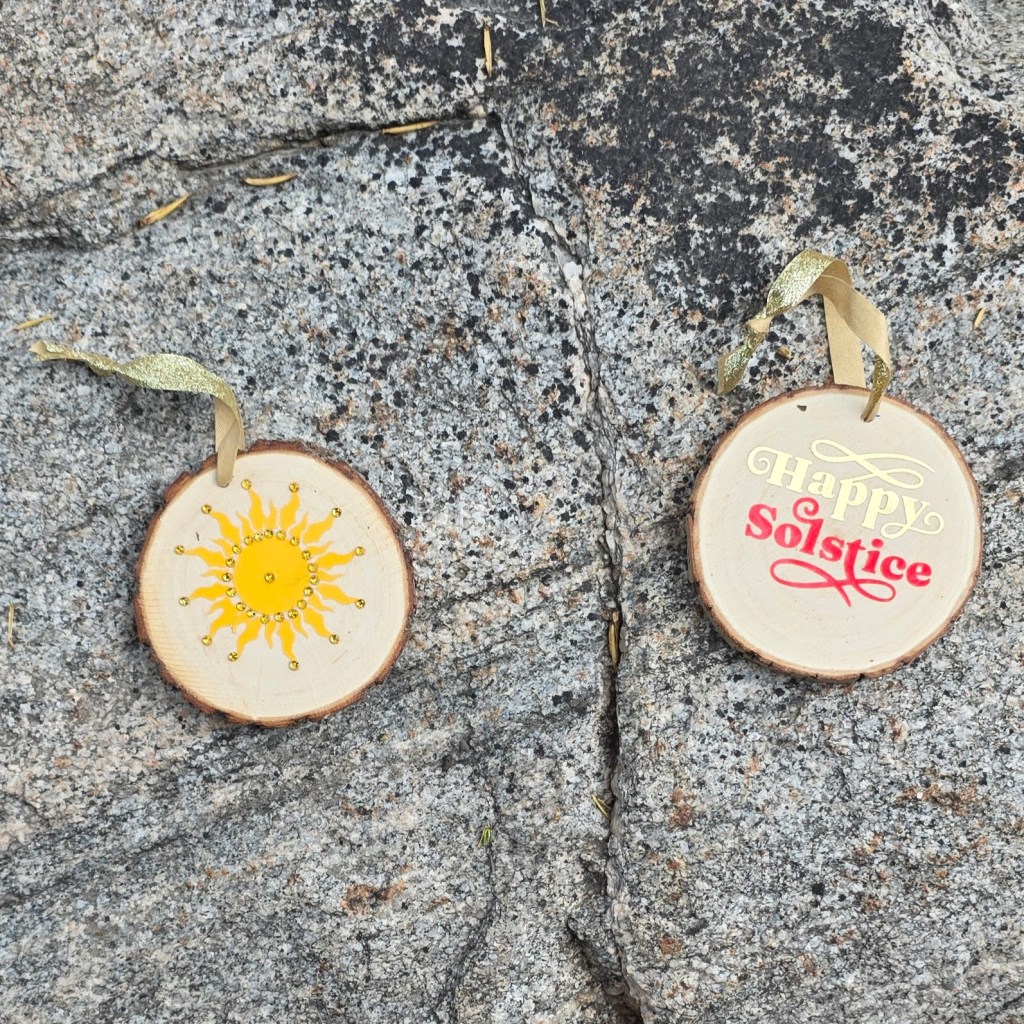 Two ornaments, the one on the left is a bejeweled sun, the one on the right says "Happy Solstice"
