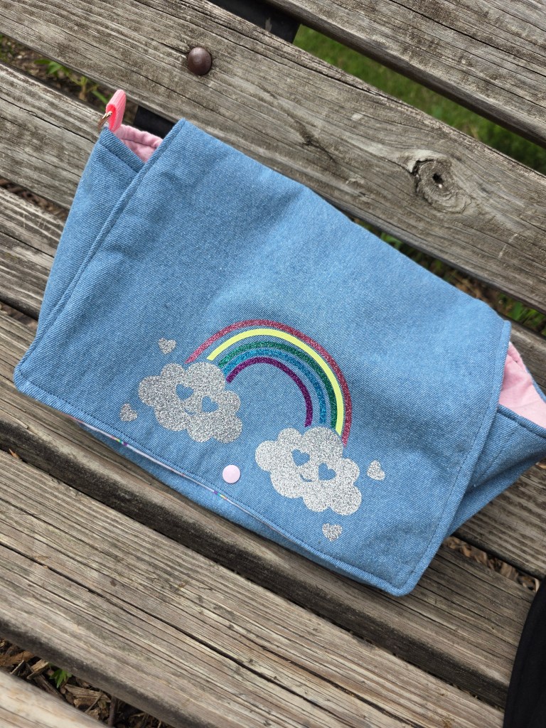a handmade, light wash denim diaper purse with a glitter vinyl rainbow with smiley heart clouds ironed on the flap. 