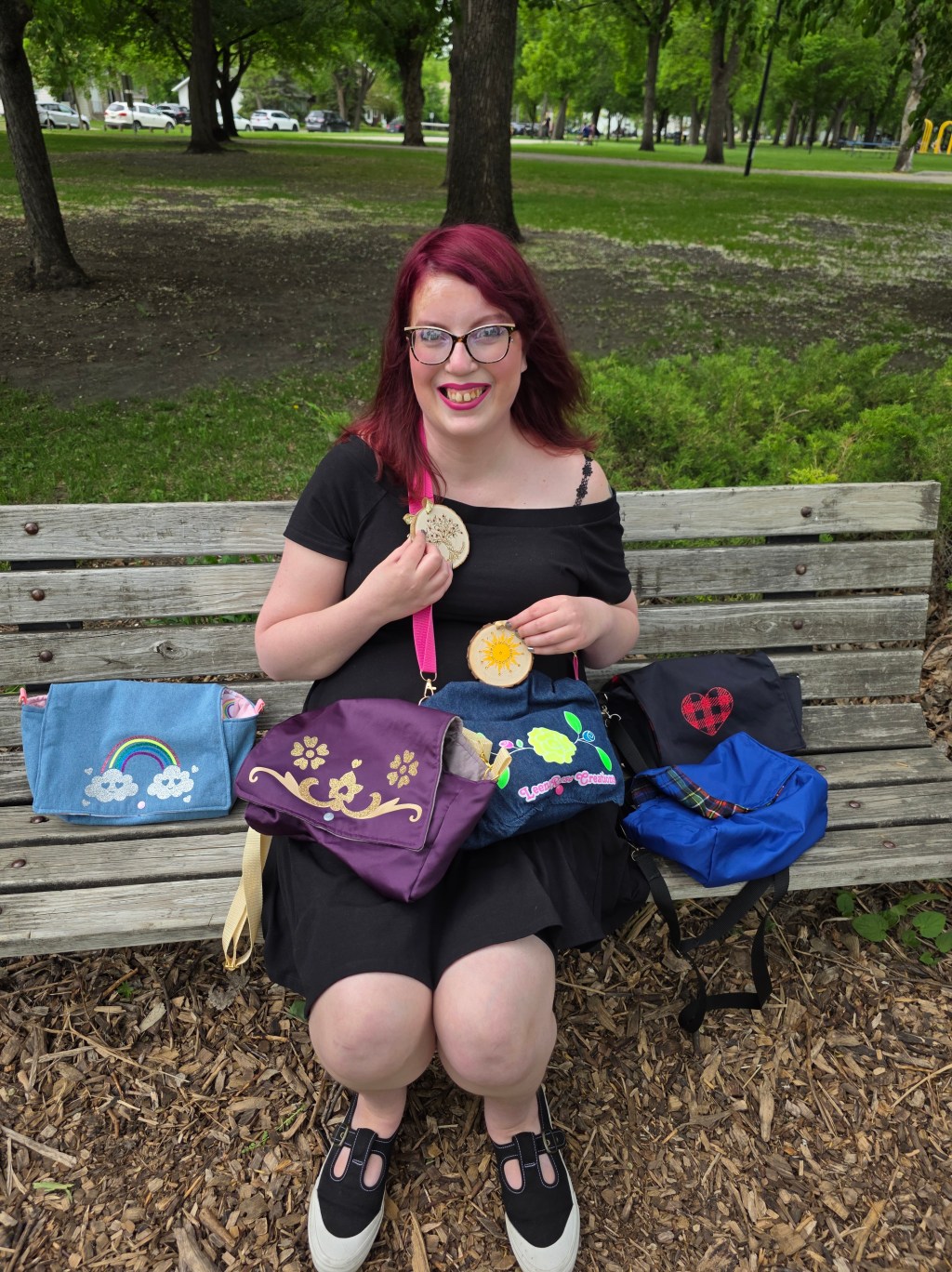 LeenaRose sitting on a University Park bench surrounded by three purses and holding two purses of her own creation. She is also holding up two wooden Winter Solstice ornaments she decorated