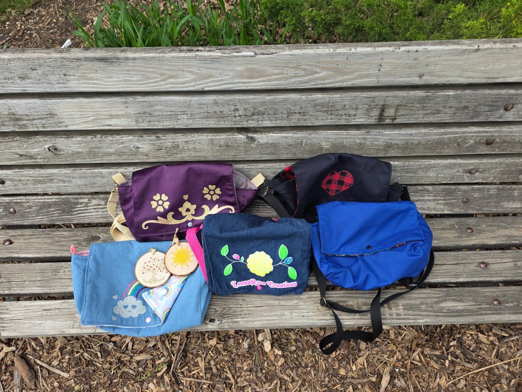 Five different diaper purses of various colors and designs and two different Winter Solstice ornaments sitting artfully on a University Park bench 